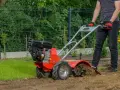 Mężczyzna w rękawiczkach prowadzi czerwoną glebogryzarkę, która spulchnia ziemię w ogrodzie.