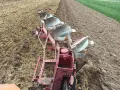 Czerwony pług z kilkoma lemieszami pracuje na polu, odwracając ziemię. Widać punkt ciągu pługa, gdzie maszyna zagłębia się w glebę.