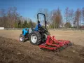 Niebieski traktor z czerwoną broną wykonuje bronowanie pola. W tle drzewa i błękitne niebo.