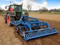 Zielony traktor z niebieskim agregatem uprawowym wykonuje podorywkę lub talerzowanie na polu.