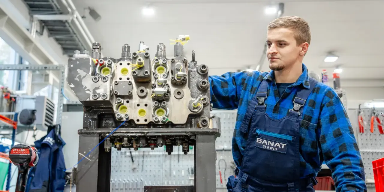 Mechanik w niebieskim kombinezonie pracuje nad budową rozdzielacza hydraulicznego w warsztacie.