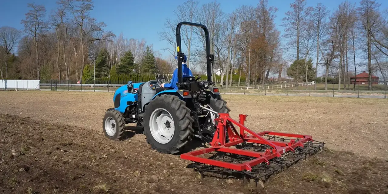 Niebieski traktor z czerwoną broną wykonuje bronowanie pola. W tle drzewa i błękitne niebo.