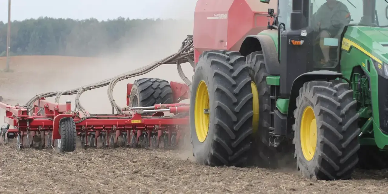 Zielony traktor z przyczepioną maszyną wykonuje wałowanie pola, podnosząc tumany kurzu.