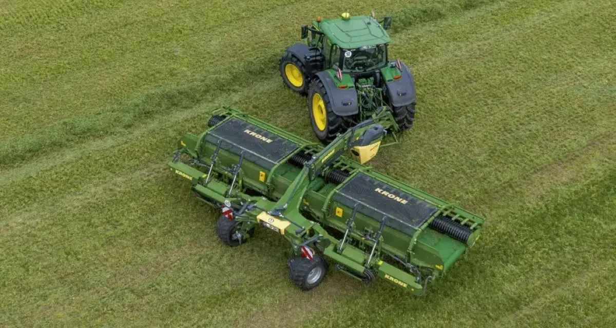 Zielony traktor z podłączoną zgrabiarką taśmową Krone pracuje na zielonym polu, zbierając skoszoną trawę.