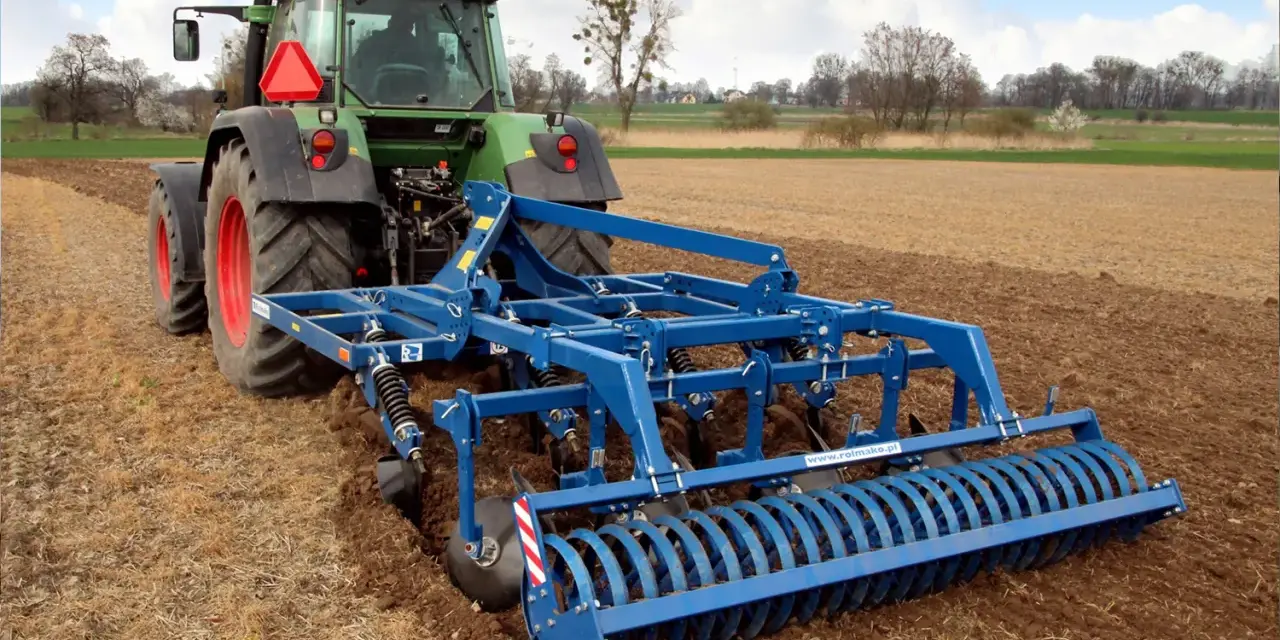 Zielony traktor z niebieskim agregatem uprawowym wykonuje podorywkę lub talerzowanie na polu.