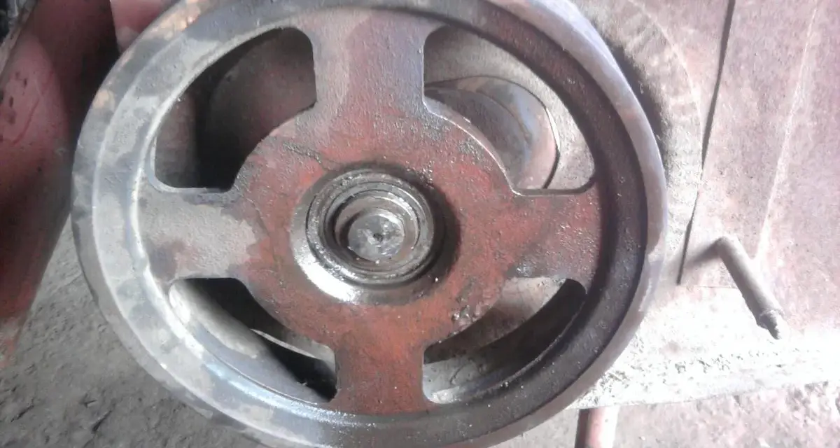 Metalowe koło pasowe kosiarki rotacyjnej, lekko zabrudzone i naoliwione, z widocznym łożyskiem pośrodku.