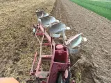 Czerwony pług z kilkoma lemieszami pracuje na polu, odwracając ziemię. Widać punkt ciągu pługa, gdzie maszyna zagłębia się w glebę.