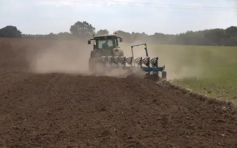 Traktor z pługiem wykonuje uprawę bezorkową, wzbijając tumany kurzu na polu.