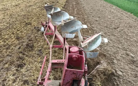 Czerwony pług z kilkoma lemieszami pracuje na polu, odwracając ziemię. Widać punkt ciągu pługa, gdzie maszyna zagłębia się w glebę.