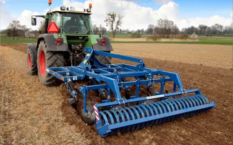Zielony traktor z niebieskim agregatem uprawowym wykonuje podorywkę lub talerzowanie na polu.