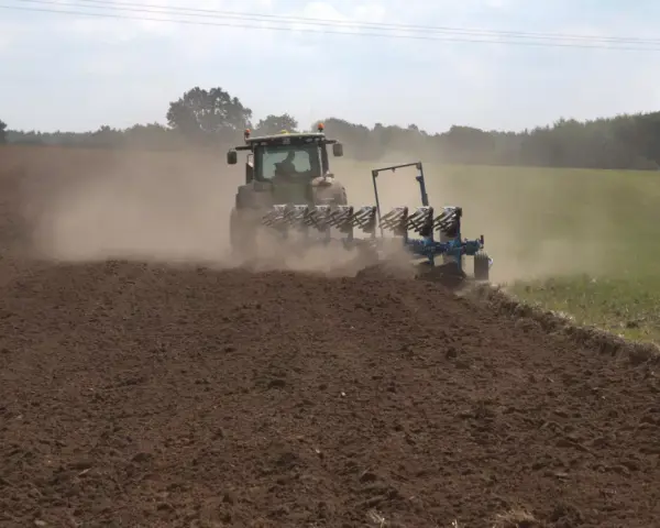 Traktor z pługiem wykonuje uprawę bezorkową, wzbijając tumany kurzu na polu.