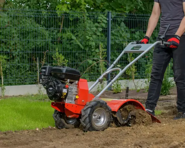 Mężczyzna w rękawiczkach prowadzi czerwoną glebogryzarkę, która spulchnia ziemię w ogrodzie.