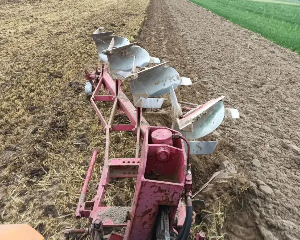 Czerwony pług z kilkoma lemieszami pracuje na polu, odwracając ziemię. Widać punkt ciągu pługa, gdzie maszyna zagłębia się w glebę.