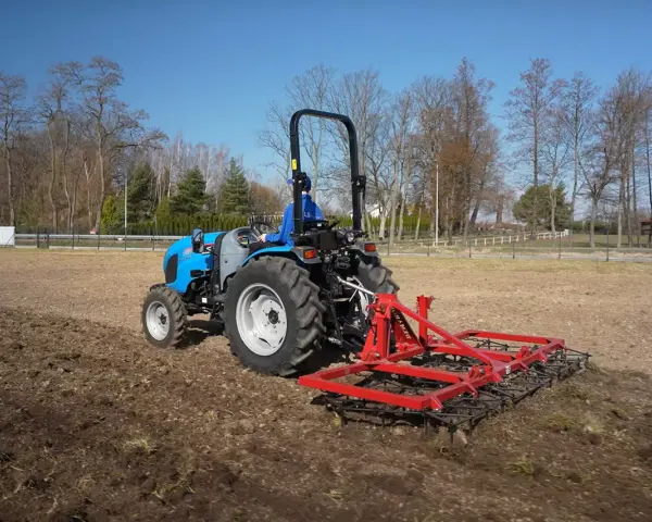 Niebieski traktor z czerwoną broną wykonuje bronowanie pola. W tle drzewa i błękitne niebo.