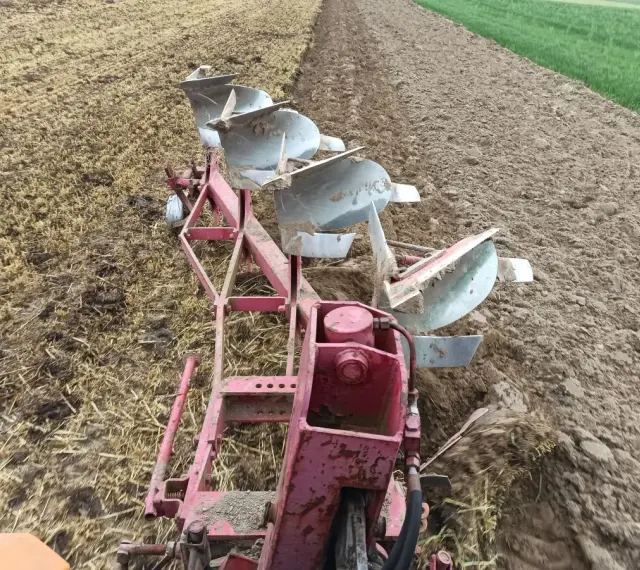 Czerwony pług z kilkoma lemieszami pracuje na polu, odwracając ziemię. Widać punkt ciągu pługa, gdzie maszyna zagłębia się w glebę.