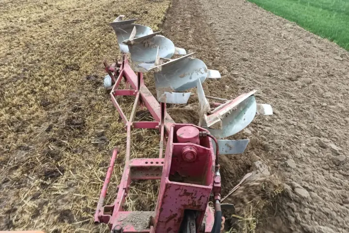 Czerwony pług z kilkoma lemieszami pracuje na polu, odwracając ziemię. Widać punkt ciągu pługa, gdzie maszyna zagłębia się w glebę.