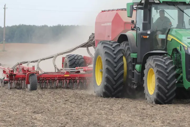 Zielony traktor z przyczepioną maszyną wykonuje wałowanie pola, podnosząc tumany kurzu.