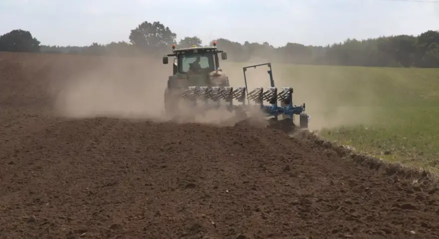 Traktor z pługiem wykonuje uprawę bezorkową, wzbijając tumany kurzu na polu.