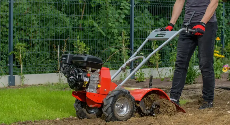 Mężczyzna w rękawiczkach prowadzi czerwoną glebogryzarkę, która spulchnia ziemię w ogrodzie.