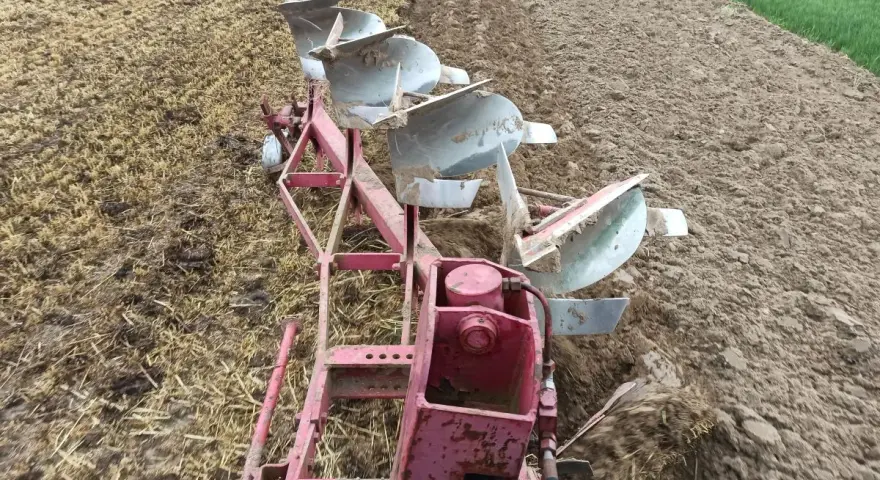 Czerwony pług z kilkoma lemieszami pracuje na polu, odwracając ziemię. Widać punkt ciągu pługa, gdzie maszyna zagłębia się w glebę.
