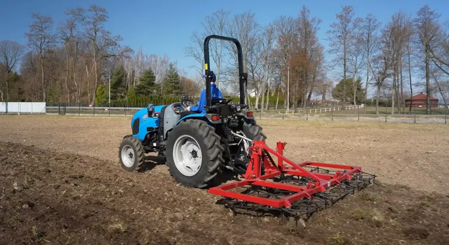 Niebieski traktor z czerwoną broną wykonuje bronowanie pola. W tle drzewa i błękitne niebo.
