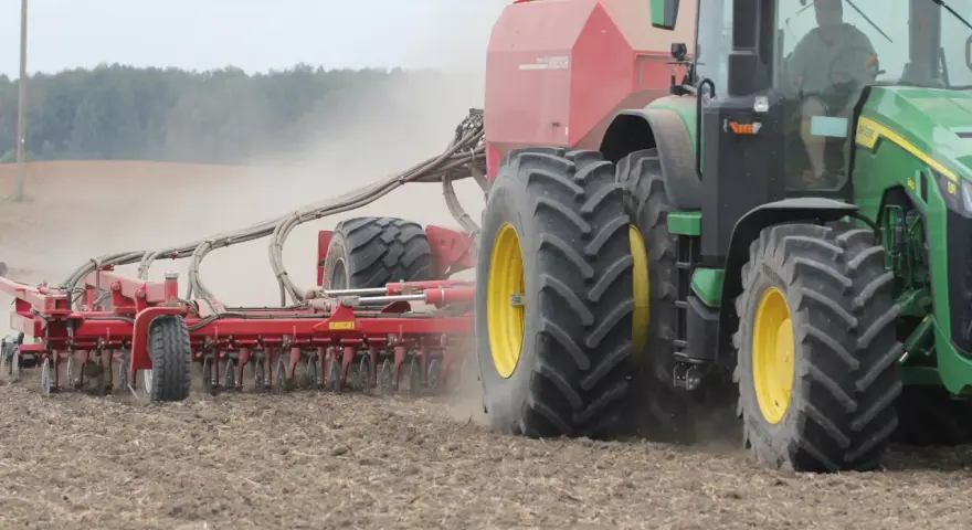 Zielony traktor z przyczepioną maszyną wykonuje wałowanie pola, podnosząc tumany kurzu.
