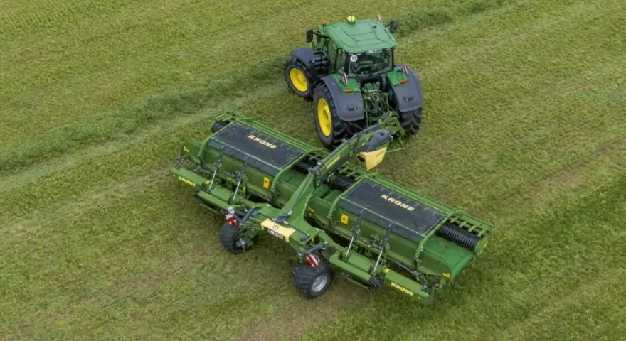 Zielony traktor z podłączoną zgrabiarką taśmową Krone pracuje na zielonym polu, zbierając skoszoną trawę.