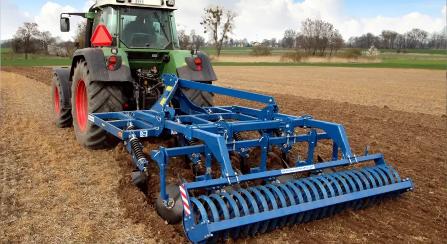 Zielony traktor z niebieskim agregatem uprawowym wykonuje podorywkę lub talerzowanie na polu.