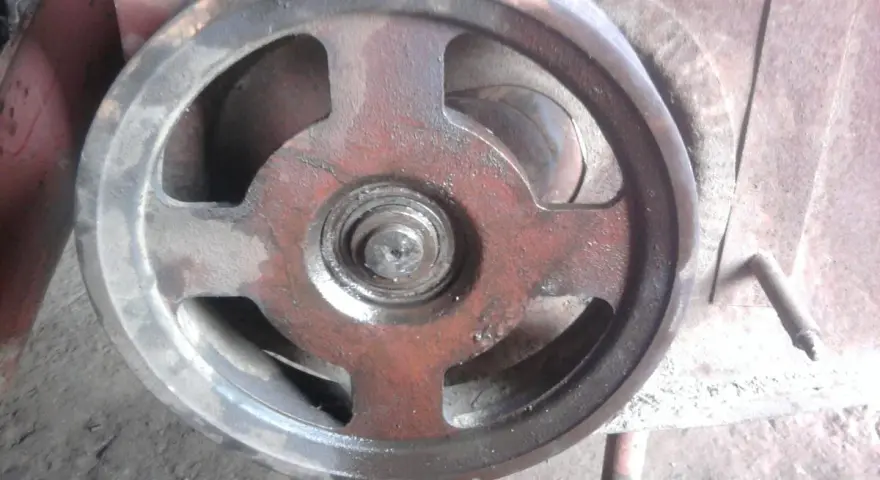 Metalowe koło pasowe kosiarki rotacyjnej, lekko zabrudzone i naoliwione, z widocznym łożyskiem pośrodku.