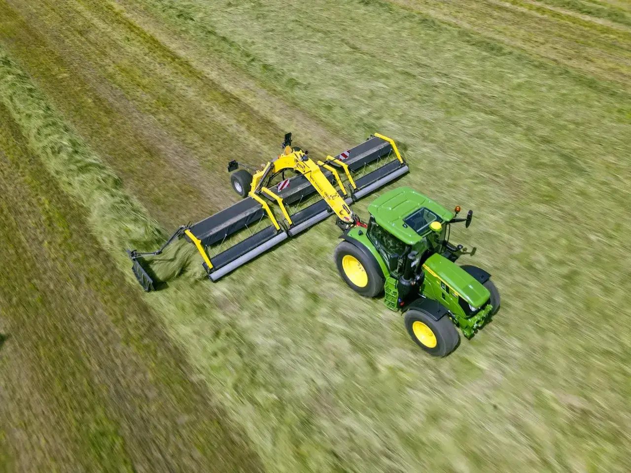 Zielony traktor z żółtą zgrabiarką taśmową pracuje na zielonym polu, zbierając skoszoną trawę.