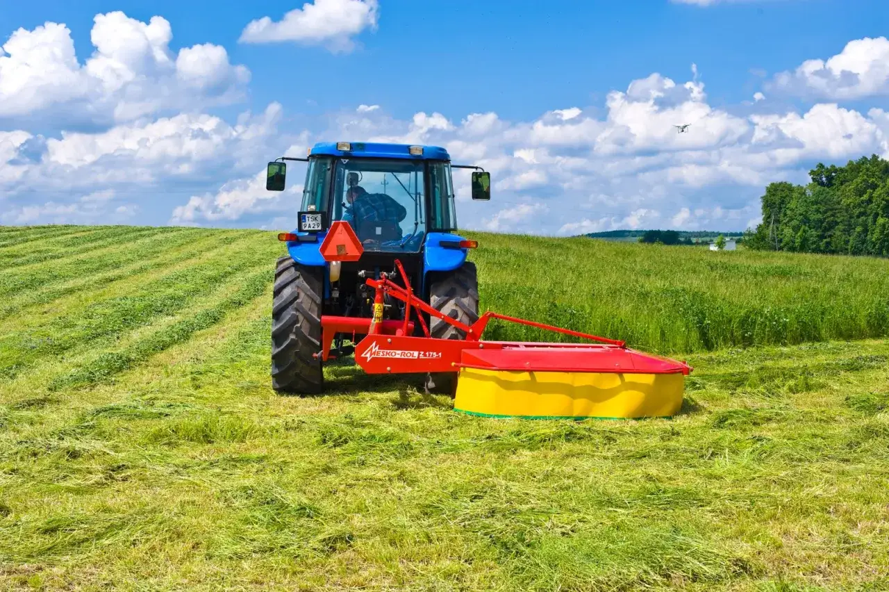 Niebieski traktor z kosiarką rotacyjną Mesko-Rol Z-175-1 pracuje na zielonym polu, tworząc równiutkie pasy skoszonej trawy.