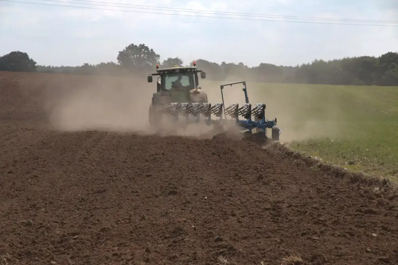 Traktor z pługiem wykonuje wykaz działań agrotechnicznych, orząc pole i wzbijając tumany kurzu.
